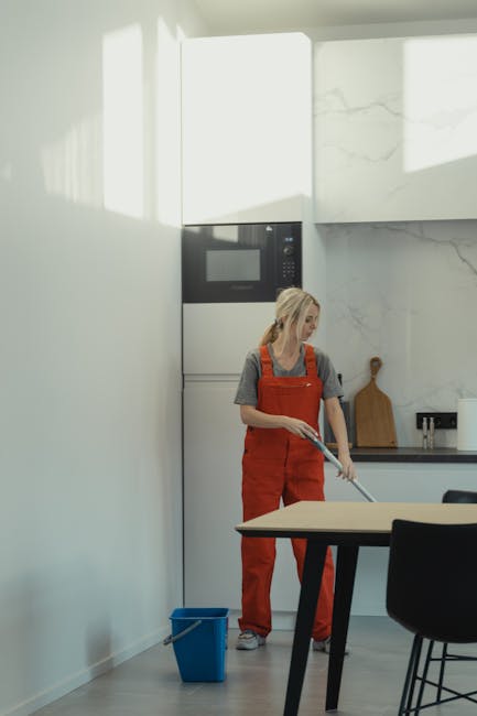 In a professional cleaning setting, a person dressed in casual attire is using a mop with a flat cleaning head to surface clean a smooth, tiled floor. The mop's handle is extended, and the person is h