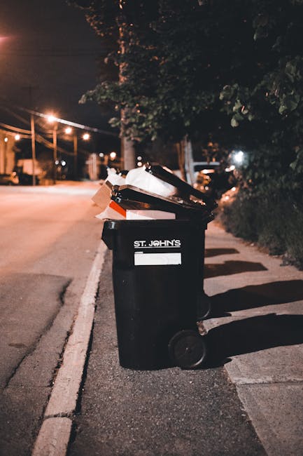 Five black wheeled waste bins with yellow lids lined up against a plain off-white wall in an outdoor area, with a concrete ground surface. The bins are made of durable plastic, each with a white label