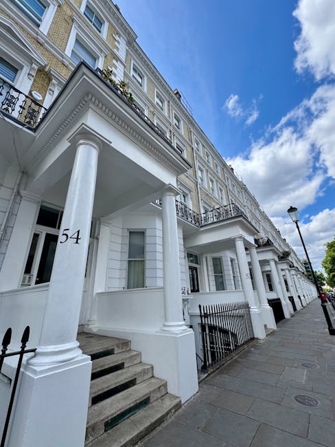 Exterior view of a row of white, multi-story residential buildings with classical architectural elements, including columns supporting covered porches at the entrance. The buildings feature large windows and black wrought iron railings along small front gardens. The sidewalk is paved with gray stones, and a black lamppost is visible beside the buildings. The sky above is partly cloudy with patches of blue, reflecting daylight that highlights the building's cleanliness and well-maintained appearance. This image exemplifies thorough exterior house cleaning and maintenance, as provided by Maida Vale Cleaners, specialists in surface cleaning and deep cleaning for residential properties in Maida Vale.