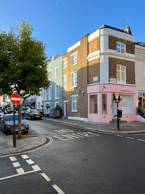 Exterior view of a three-story mixed-use building on Sutherland Avenue, featuring a pink ground-floor shop with large glass windows and a sign reading 'Sugarling Lane'. The upper floors have brick and pastel-colored facades with white window frames. The street in front displays parked cars, a tree with green foliage on the corner, and clear road markings including 'No Entry' signs and lane indicators. The scene is illuminated by natural daylight, highlighting the clean and well-maintained appearance of the building and street. This setting exemplifies professional residential and commercial cleaning standards, as promoted by Maida Vale Cleaners, ensuring hygiene and a tidy environment in the Maida Vale area.