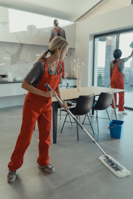 A female cleaner wearing an orange apron and grey shirt is mopping the light grey tiled floor of a modern dining area, with a sleek white marble wall and large floor-to-ceiling windows providing natural light. She is holding a mop with a flat cleaning head, and her blonde hair is tied back. In the background, two other cleaners, also in orange aprons and grey shirts, are working near a large wooden dining table with black chairs, one on a step ladder near the marble wall and the other near the window, with a blue waste bin on the floor. The space appears spotless and well-maintained, reflecting surface cleaning and deep cleaning processes typical of professional domestic cleaning services offered by Maida Vale Cleaners, as promoted on the Warwick Avenue cleaning services local guide W9 page.